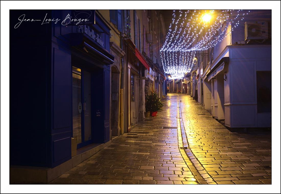 A narrow, old-fashioned lane unfolds toward a distant vanishing point, its cobblestones slick with recent rain and catching the glow of street lighting. A warm, golden lamp hangs above and spills a pool of amber across the pavement, creating a bright ribbon that runs down the center and reflects in the shallow grooves between the stones. Overhead, a canopy of cool white fairy lights is strung across the street, their small points like a constellation that softens the night and adds a festive, intimate atmosphere.
Shuttered shopfronts line both sides of the alley: deep blues and muted neutrals form a quiet palette, with window frames and recessed doorways stepping back into shadow. A single potted conifer—maybe a small Christmas tree—stands near one doorway, lending a touch of green and a domestic note to the otherwise empty thoroughfare. The buildings are close and low, their facades textured and slightly worn, suggesting an old neighborhood with character.
The surface of the street is the scene’s star: wet, reflective, and laid out in neat, rectangular stones that guide the eye forward. A narrow channel or line of stones runs down the center, accentuating perspective and rhythm. The contrast between the cool blue tones in the shadows and the warm yellows where the lamp hits gives the composition a cinematic feel—quiet, slightly melancholic, and beautiful in its stillness.
