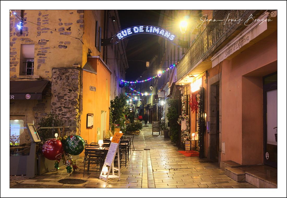 A narrow, cobblestone alleyway at night is bathed in warm lamplight and the colored glow of festive string lights. The street slopes gently away, drawing the eye toward a bright arched sign that proclaims the lane’s name and a pool of light at the far end. The wet stones of the pavement pick up reflections from the lamps and decorations, giving the ground a soft, almost liquid sheen.
On either side, low shopfronts and cafés press close together — ochre and rosy plaster facades, patches of exposed stone, a small wrought-iron balcony overhead. A handful of bistro tables and chairs sit quietly outside, accompanied by a sandwich-board menu and potted evergreens. Christmas touches — oversized glossy baubles, wreaths and garlands, and little decorated firs — punctuate the scene, suggesting holiday cheer without crowd or bustle.
The lighting is intimate and layered: a golden streetlamp throws a halo across the passage, while strings of blue and purple bulbs stitch the rooftops together in a festive canopy. The composition has a cozy, cinematic quality — textures of stone and wood, the play of shadow and color, and a sense of calm anticipation as if the street is holding its breath between quiet evenings and the arrival of visitors.

