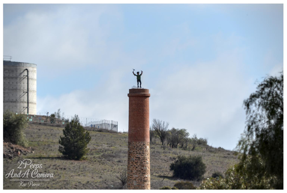 Photograph showing a tall, historic red brick and stone chimney stack on a dry, grassy hillside in Burra, South Australia.

A statue of a figure with arms raised is positioned triumphantly at the top. A modern cement silo is visible on the left in the background under a partly cloudy sky.