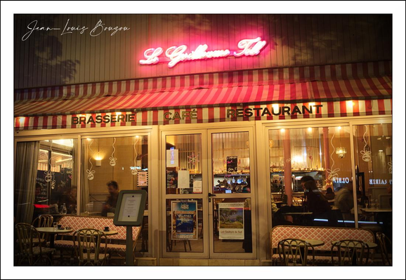 A small, warmly lit brasserie at night fills the frame, its striped red-and-white awning announcing a familiar, old-world charm. Above the awning a neon cursive sign glows pink, throwing a soft halo over the façade. The word “BRASSERIE” and “RESTAURANT” are stenciled across the front, and a freestanding menu board sits beside the glass door like an invitation.
Through the large windows you can see the golden interior light spilling onto the sidewalk, silhouettes of diners and waitstaff moving in the cozy space. Round bistro tables with woven chairs and red-checked tablecloths are arranged on the narrow pavement, small enough to suggest intimate conversations over wine. Reflections and warm highlights dance on the glass, while posters and small notices are taped to the door, lending the place a lived-in, neighborhood feel.
The scene breathes a quiet, late-evening atmosphere — the kind of place that promises comfort and familiarity: clinking cutlery, low conversation, the smell of frying onions and roasted garlic just out of sight. It feels like a small refuge from the street, a pocket of soft light and humanity against the night.

