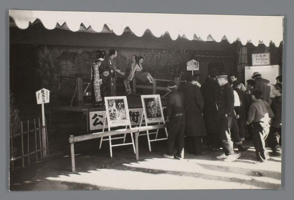 The image depicts a historical scene at what appears to be an outdoor marketplace or festival in Japan. Several individuals are gathered around display boards featuring artwork, possibly traditional Japanese prints known as ukiyo-e. A few people wearing Western-style suits and hats stand in line or observe the displays, while others wear kimonos indicative of cultural attire from that era.

There's a structure with signage where one person seems to be handling transactions or admissions for an event labeled "公団菊展," which translates to something like "Public Industrial Exhibition." This suggests some formality or organized activity taking place. The overall mood is casual, yet there are indications of cultural and social interactions occurring.

The photograph has a grainy texture typical of black-and-white film from the mid-20th century. It captures everyday life with an unposed quality, giving it authenticity as if documenting a moment in time rather than staging for viewership.