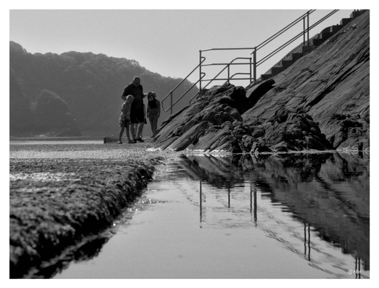 A man and his two sons stand at the end of a rough-topped sea wall. The three are 'crabbing'. Man-made steps hewn into the natural rockface, with metal guard rail to protect people from the steep sides. The rockface and rail are reflected in the water.