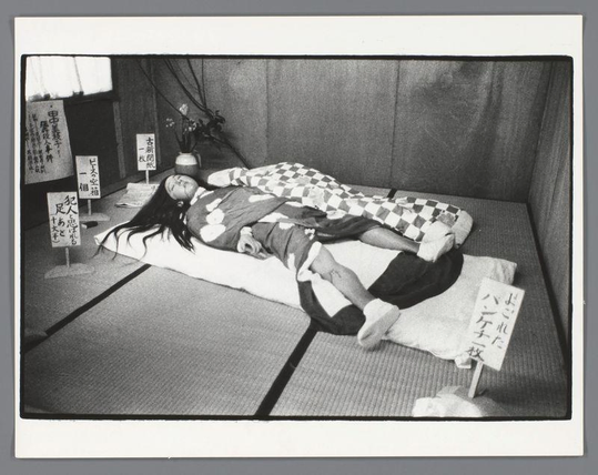 The image depicts a black and white photograph of an individual lying on the floor, seemingly unconscious or asleep. The person appears to be wearing dark clothing with some lighter-colored patches, possibly denim shorts. They are sprawled out across two traditional Japanese tatami mats laid side by side, which suggest this scene is set in Japan.

In the background, there's a plain wall and what looks like an indoor setting with minimal decoration or furnishings visible. There are several signs around the individual on small posts that contain text written in Japanese characters. These signs seem to be placed at different distances from the person, some closer by their head and others further away near one of the corners.

The photograph is taken in a manner reminiscent of documentary photography, capturing an unembellished moment with stark lighting and high contrast between light and shadow areas. The composition focuses on the individual's face down to mid-torso level, emphasizing their state or condition without any explicit contextual information provided within the image itself.