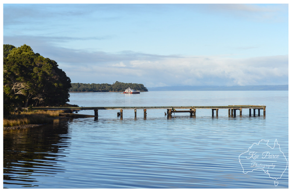 A long, low wooden jetty extends into a vast, calm blue water body. On the horizon, a cruise or ferry boat is visible, flanked by forested land on the left. The sky is bright blue with scattered white clouds.