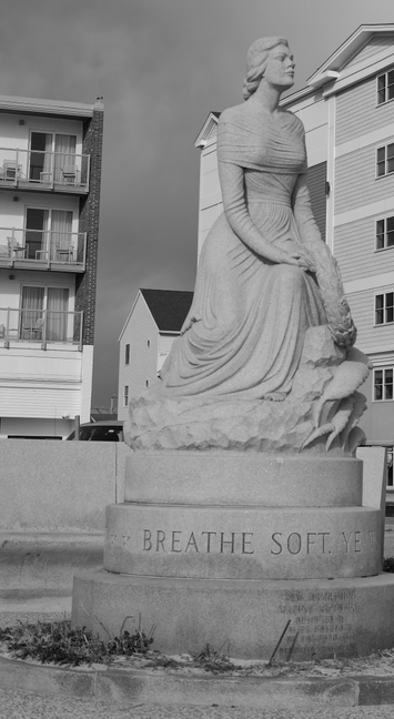 Statue of a woman with a bare shoulder dress that breaks into rufflers at her feet on a round pedestal with hotel buildings in the background