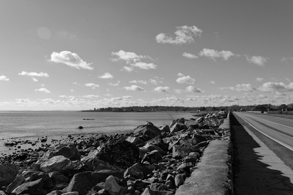 Black and white image:  to the far right there's a road,  then a concrete wall and a large mass of rocks meeting the ocean,  in the background a segment of coastline bends towards the left.