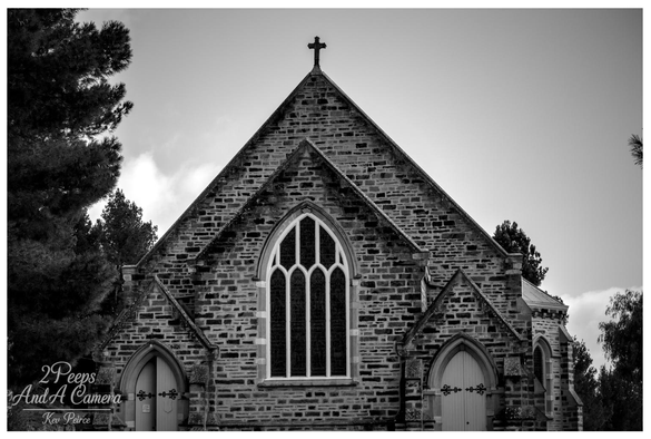 Black and white photograph capturing a frontal, symmetrical view of a historic stone church's facade.

Features include a large Gothic style arched window with white tracery, two smaller side entrances, and a cross atop the main gable. Dark trees frame the structure on the left against a bright sky.