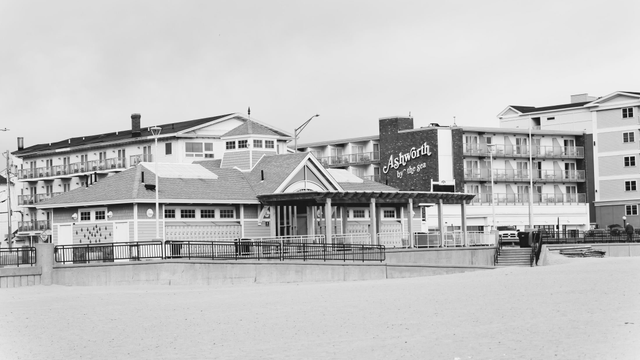 The bottom 1/3 is a sandy beach out of which a rambling hotel building rises with a sign on one wall that says Ashworth by the Sea with a featureless grey sky above
