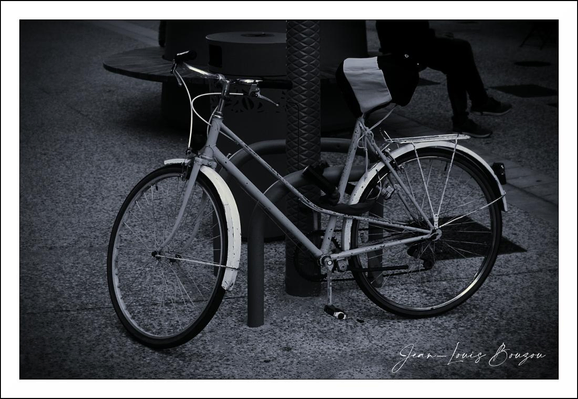 A single bicycle is locked to a stout metal post, its classic step-through frame hunched forward as if pausing mid-journey. The photo’s cool, desaturated tones turn the scene almost monochrome, emphasizing the pale, curved fenders that rim both wheels and the gleam of metal spokes against the dark pavement. The front wheel is slightly turned, the handlebars angled toward the viewer, and a narrow saddle and rear luggage rack give the bike a utilitarian, well-used character. Soft highlights pick out the chain, the textured grips, and the subtle scratches along the frame; shadows pool beneath the tires and stretch away across the concrete. In the background a bench sits in shadow; a seated figure is only partly visible, reduced to a silhouette and a suggestion of legs, which adds a quiet human presence without drawing focus from the bicycle. The overall mood is calm and contemplative, as if the street has been held in a single, hushed moment between departures. 