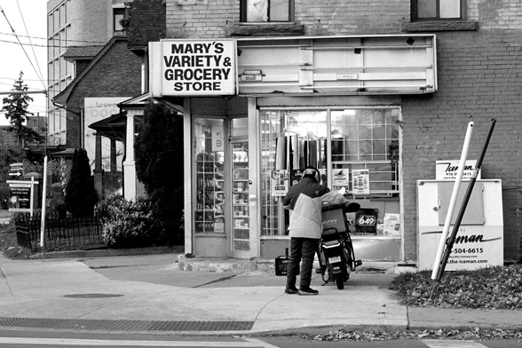 In the middle of the background is a corner store called Mary’s Variety and Grocery Store. In the left background are the buildings further up Broadview. To the right of the store is an ice machine, slightly tilted on uneven ground. Just right of centre is a food courier standing next to his bike.