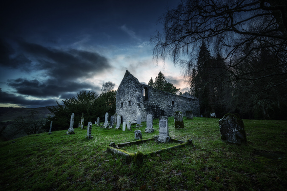Die Steinruine der Alten Pfarrkirche in Blair Atholl, Schottland, dominiert einen huuegeligen Friedhof mit alten Grabsteinen. Eine dramatische Daemmerungsstimmung mit dunklen Wolken und einem letzten Lichtstreifen am Horizont umgibt die Szene, waehrend kahle Baeume den rechten Bildrand rahmen. Die Weitwinkelaufnahme betont die Weite des Gelaendes und die melancholische Schoenheit der historischen Staette.