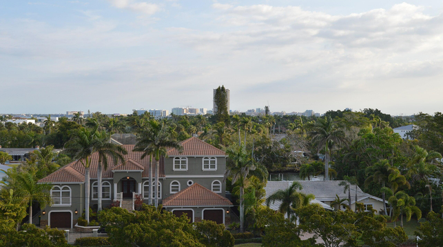 A photo overlooking houses surrounded by palm trees. There's a prominent mansion in the foreground. Larger buildings can be seen in the distance. The sky is a hazy blue and filled with white clouds.