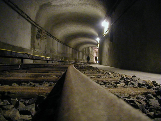 the picture (taken from the rail directly) shows a view along the tube of the then closed S-Bahn line. It takes a wide turn to the right. There are two people seen wandering off to the end of the tunnel. On the right wall to lit lamps emitting cold white light are seen. The trails are kind of rusty.