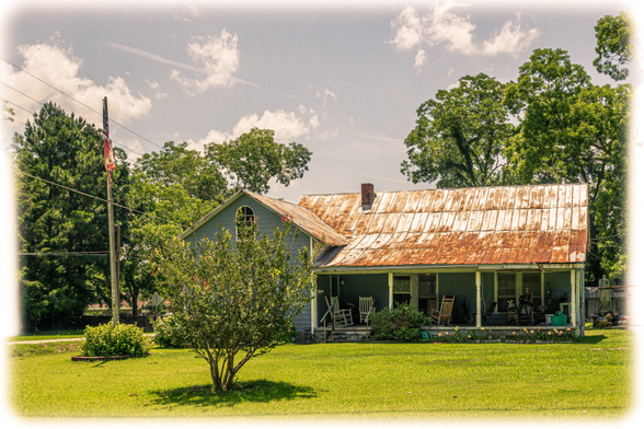 A weathered single-story house with sage green siding and a heavily rusted corrugated metal roof showing orange-brown discoloration and deterioration. The house features a gabled front section with a decorative arched window and a long covered front porch spanning the length of the structure. Several rocking chairs sit on the porch. A red brick chimney rises from the center of the roof. The front yard has well-maintained green grass with a small ornamental shrub or tree in the foreground. Mature trees with full green canopies frame the house on both sides and in the background. A utility pole with power lines and what appears to be an American flag is visible on the left side. The image has a soft vignette border effect and is photographed under partly cloudy skies.