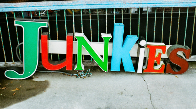 A  color photo of a light-up sign that says 'Junkies' sitting on the ground outside a bric-a-brac shop.