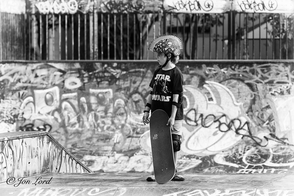 This is a black and white street photo in landscape format of a boy holding his skateboard and wearing a scary safety helmet. Somerset, Singapore (2017).

In the centre of the image is a boy of, say 10 years old, viewed nearly in profile and facing to the left with his front and left side in view. He is standing in a urban skateboard park and not surprisingly is holding a skateboard in his left hand with the base of the board resting on the ground. Our young man is wearing trainers, shorts, a T-shirt with 'Star Wars' on the front and dark glasses. He has come prepared and has knee pads, elbow pads and a fantastic safety helmet that starts at the forehead covers the head, around the top of the ears and finishes at the top of the neck. In the middle of the helmet, stretching from the front to the rear are a number of parallel rows of reptilian or Mohawk like spines, plastic or rubber I hope. The helmet is black but with numerous white insignia of skulls, lightning bolts and crossed guitars etc. 
In the background are ramps and railing all with grafetti spray painted on all the surfaces. 
A young man not to be trifled with !