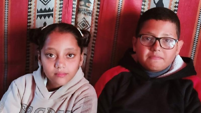 Two children, a girl (Aylan) with hair in buns and a boy (Elias) in glasses, sit side-by-side, facing the camera, against a red and patterned backdrop.
