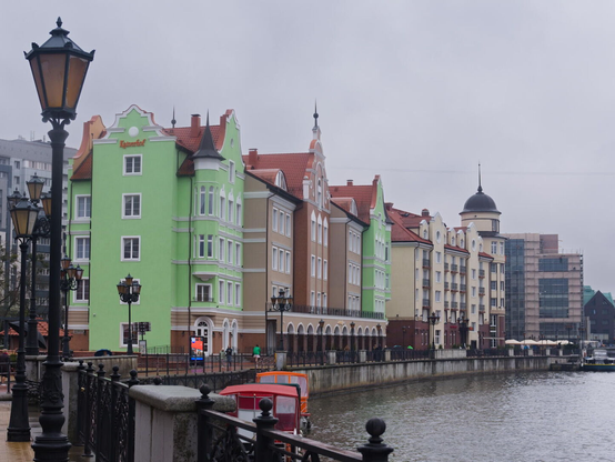 The image captures a vibrant and picturesque riverside promenade. The viewer's perspective is from the water level on one side of the river, looking up towards the cityscape across the way. The architecture along the waterfront features colorful buildings with ornate facades that add to the charm of this historical district. The buildings are painted in various pastel shades, including green, beige, brown and yellow, creating a vibrant and lively atmosphere.

A few people can be seen walking along the promenade. The overcast sky lends a soft light to the scene without casting harsh shadows on the buildings or pedestrians below. There are numerous lampposts lining both sides of the promenade. A few boats can be seen docked along the riverbank. The green building on the left side stands out due to its distinct color and traditional architectural style.