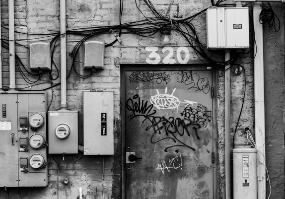 A black and white photo of the back entrance to an urban building with electrical boxes on the wall and graffiti on the door.