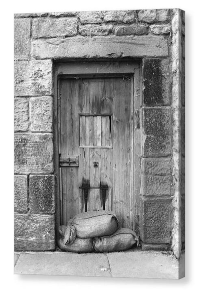 Black and white portrait photo of a small wooden door with sandbags piled in front.  The image is shown printed upon a block canvas.