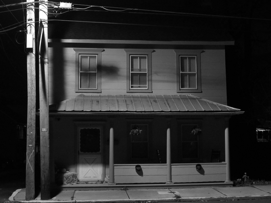 A rather unremarkable house on the corner of Center and Spruce in the town where I live. Shot during the dark early morning hours, I think I photographed it because I found the light a street lamp was casting upon it, interesting. The light fixture is out of frame and is attached to one of two utility poles on the corner of the sidewalk in front of the house which also bears a number of electrical cables. There are three windows on the second floor, below which is a slanted tin porch roof, and all lit by the street lamp. The door to the place is on the left and two windows on this first floor are to the right of it. The porch below the roof is in shadow, except for the bottom part of the door which is lit by the street lamp.There are three columns supporting the porch roof where a solid fence is attached to them and which separates the house from the sidewalk in front of it. Two hanging plants and two potted plants adorn the porch. A hay bale with a small pumpkin sitting atop it, just to the left of the door, serve as seasonal decoration. It all adds up to a sort of stark little scene that might appear in a 1950's noir crime film. In the film something nefarious is, is about to, or has just happened inside the house and the camera is just about to show you. The music is tense and dissonant as the camera searches through several rooms until...