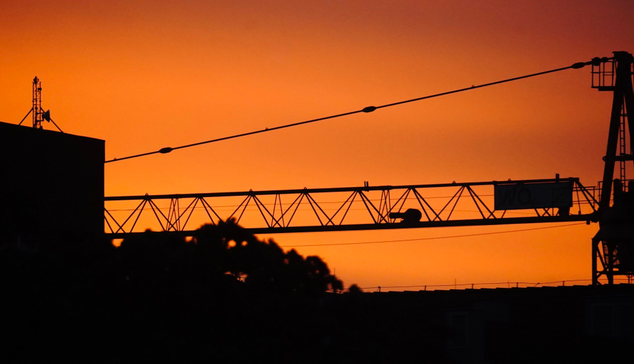 A partial close-up photo of a large crane during sunset. Due to the light conditions, only the contours of the crane are visible against the orange (fire-colored) sky. There is also part of a building on the left side, and again, we can only see its contours; the details are drowned in black.
