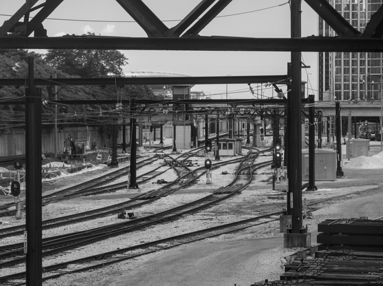 Black and white, high contrast photo emphasizing the curves of the railroad tracks and structures of the steel scaffolding that hold the overhead lines for electric trains.