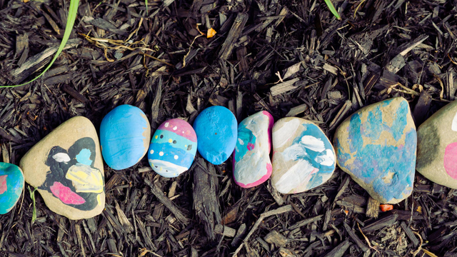 9 variously shaped and painted rocks in a row as if they were making a snake on top of brown mulch