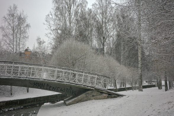 The photo shows a winter landscape with a bridge covered with snow. There are trees around, also covered with snow, and a building in the distance. Snowfall creates an atmosphere of tranquility and privacy.