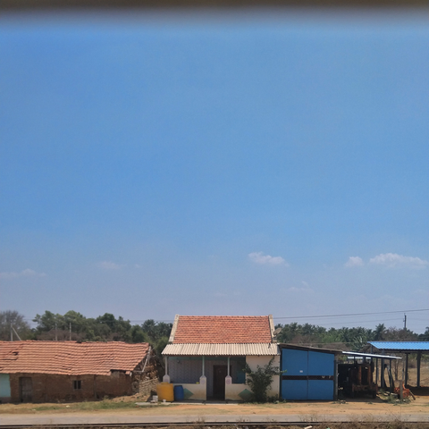 Small rural house with a red-tiled roof and a blue shed nearby, set against a clear blue sky. Sparse vegetation adds a rustic feel.