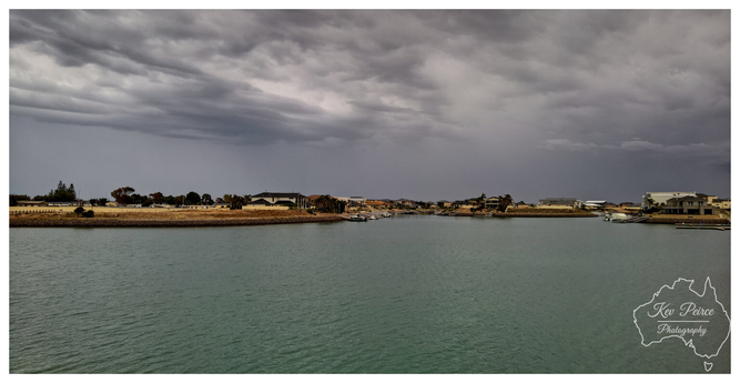 A wide, atmospheric photo of a calm water way, possibly an artificial canal or marina, lined by modern houses and properties on the distant banks.  The sky is completely dominated by thick, turbulent, dark gray storm clouds, creating a moody and dramatic contrast with the flat, blue green water.