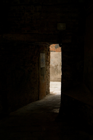Photo looking through a deeply shadowed passage at an angle, it either narrows towards the end or zig-zags, as a dark corner cuts off some of the view out from the end. Most of the image is dark, in the middle the passage opens into a narrow sunlit alley, with solid grey-white plaster under just-visible cracking red plaster on the wall opposite. The light spills in onto the grey flagstone floor and bare brick wall slanting leftwards. On that wall is a pale rectangle, maybe a sign or a plate blocking up hole, covered in graffiti.