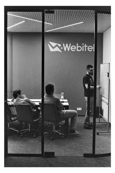 A grainy black and white photograph of an office meeting seen through a glass partition. On the right, a bearded man stands writing on a whiteboard. On the left, two men sit at a desk listening. The background wall features a large "Webitel" logo. The lighting is soft and natural