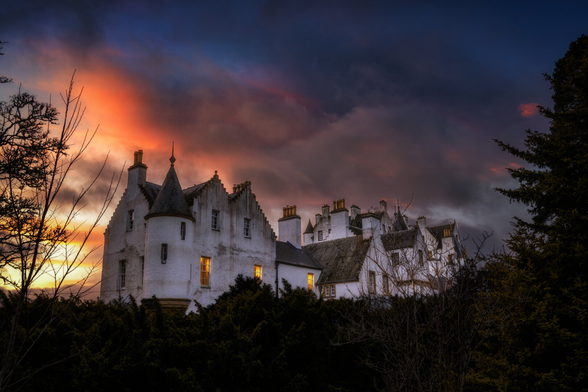 Ein historisches Gebäude mit weissen Fassaden und einem Turm ragt unter einem dramatischen Sonnenuntergangshimmel hervor. Warme Lichter in den Fenstern kontrastieren mit den dunklen Silhouetten der Bäume im Vordergrund. Die Komposition betont die architektonischen Details und die stimmungsvolle Abendbeleuchtung.