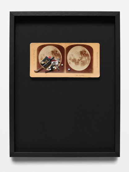 A framed artwork featuring a vintage stereoscopic card with two sepia-toned images of the full moon. Small pieces of metallic and plastic debris such as screws, clips, a soap and a magazine are arranged on the left moon image, resembling objects left behind on the moon during the apollo missions. The card is mounted on a large black background within a simple black frame.