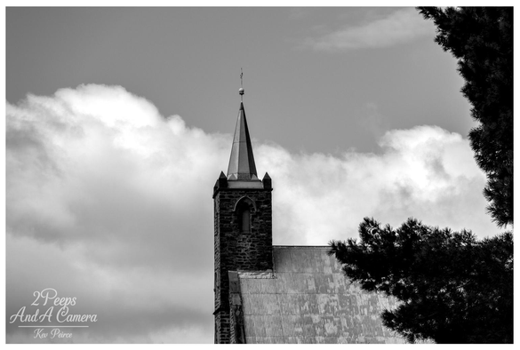 Black and white photograph showing the stone steeple and metallic spire of a Burra church, framed on the right by the dark, textured branches of a tree.

The church's corrugated iron roof is visible below the steeple against a sky filled with bright, contrasting clouds.
