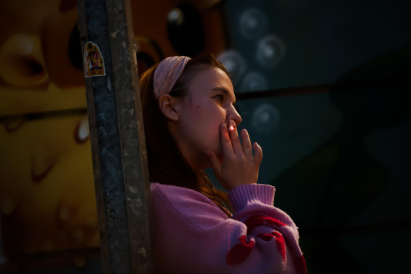 Colorful night-time portrait photo of a young woman smoking a cigarette, wearing a pink sweater, leaning against a metal pole, with an abstract colorful background.