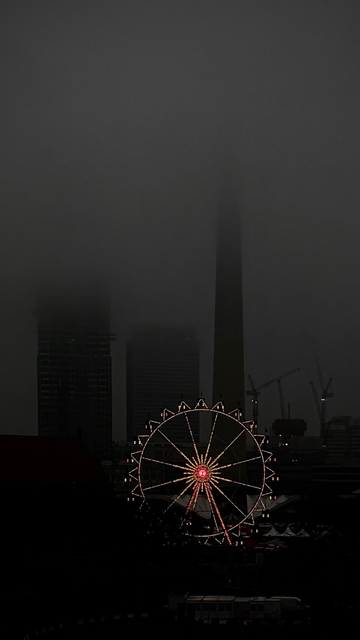 A foggy urban skyline featuring a illuminated Ferris wheel in the foreground, with tall buildings and a tower obscured by mist in the background.