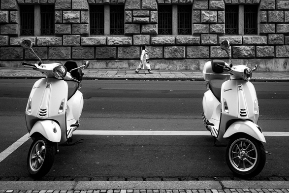The image is a black and white street scene featuring two white Vespa scooters parked on a road. The scooters are positioned symmetrically, facing the viewer. The scooter on the left has a dark wheel, while the one on the right has a light-colored wheel.

In the background, a large, imposing stone building dominates the scene. The building's facade is constructed from large, rectangular blocks, and it features several windows with ornate iron bars. A person is walking across the road in the distance, between the scooters, heading towards the building. They are wearing light-colored clothing.