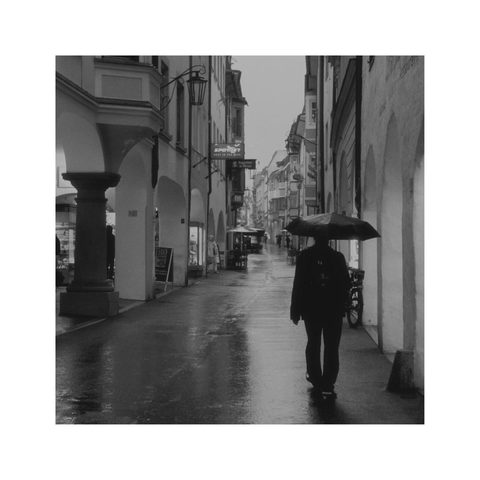 a square black and white image of a pedestrian street. it's raining, there's just one dark figure with a black umbrella walking down the deserted street