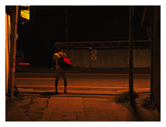A person wearing a striped shirt and shorts, carrying a prominent red and black duffel bag on their back, stands on a cracked sidewalk facing a dark, empty road. The sidewalk and asphalt are illuminated with an intense orange glow, likely from nearby streetlamps or signs. To the left, a bright yellow sign advertises "WHITE RICE," while a red streak of light-trail crosses the road, suggesting a vehicle has just passed. The background consists of a dark structure, possibly a closed establishment, with a gated or paneled facade. - Google Gemini Latest