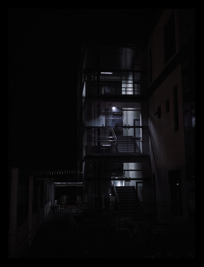 A modern building facade. The central feature is a multi-level, glass-enclosed stairwell, brightly illuminated from within, standing out sharply against the deep black surroundings. - Google Gemini Latest