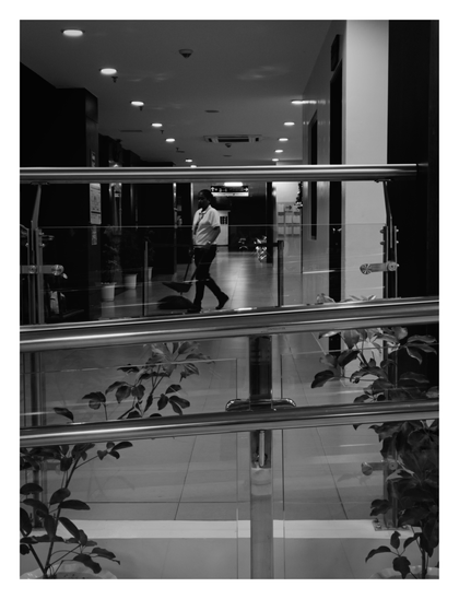 A hallway stretches into the background, illuminated by recessed ceiling lights. In the midground, a worker (likely maintenance staff, holding a broom/dustpan) walks from left to right. The foreground is dominated by the horizontal lines of a stainless steel and glass railing, which partially obscures potted plants near the floor. - Google Gemini Latest
