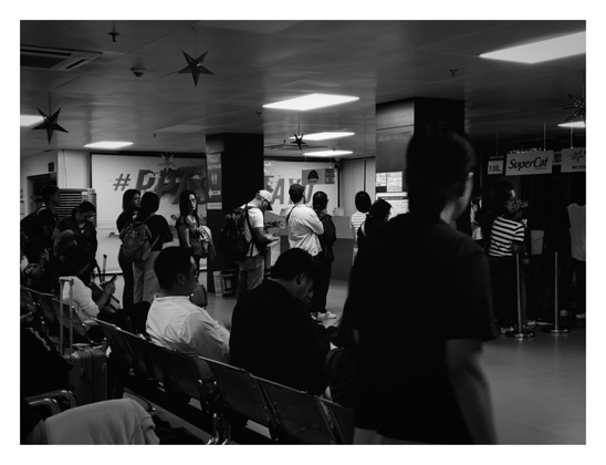 A crowded indoor waiting area, likely a ferry or transport terminal. Passengers are seen sitting on metal benches or standing in groups, surrounded by luggage and background signage, including "SuperCat" and "Public Restroom." A blurred figure in the immediate foreground frames the depth of the busy scene. - Google Gemini Latest