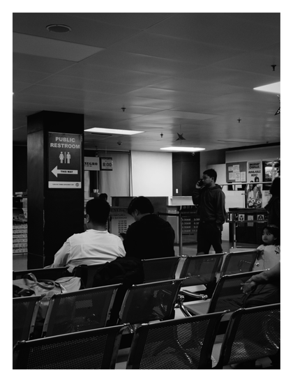 A busy transportation waiting area, likely a port or bus terminal. Rows of perforated metal seating fill the foreground, occupied by several travelers, including two men seen from behind and a small child seated on the right. In the center-right, a young man walks past while talking on a mobile phone. A prominent sign on a pillar directs travelers to the "PUBLIC RESTROOM." Departure schedules and a "Mister Donut" advertisement are visible in the background, beneath the illuminated drop ceiling. - Google Gemini Latest
