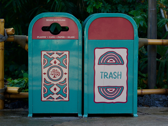Two adjacent themed trash cans in Adventureland. Both are teal with red accents and cream panels featuring bold geometric designs. The left bin is labeled “Mixed Recycling” with a circular opening; the right reads “Trash” with stylized arc patterns.