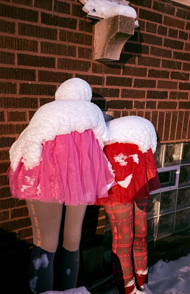 Two lower-body mannequins stand outside against a brick wall, their backs to the camera, covered in fresh snow. One wears gray leggings and a bright pink tulle skirt, while the other wears red patterned leggings and a red skirt, creating a humorous scene where the piled snow resembles fluffy tops or heads on the figures.
