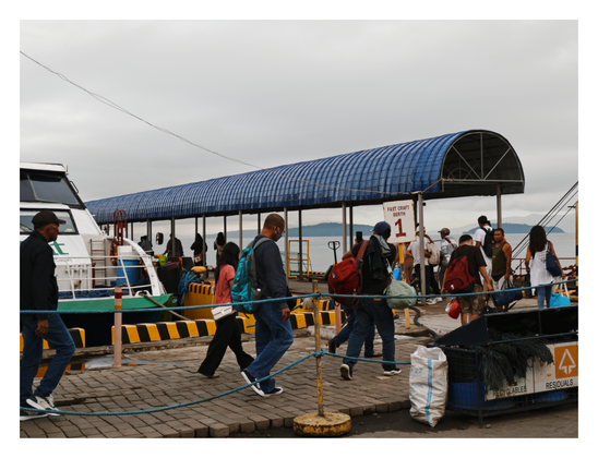 Several passengers walking along a paved pier under a long, arched blue metal roof. A sign clearly indicates "FAST CRAFT BERTH 1." A fast craft ferry is docked on the left, and travelers are seen carrying bags and backpacks. Distant hills rise from the sea under a gray, overcast sky. - Google Gemini Latest
