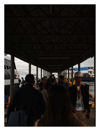 A crowd of people walking through a long, shadowed, covered terminal walkway (gangway) towards a large docked ferry and the bright, overcast harbor area. - Google Gemini Latest
