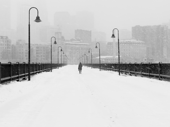 A black and white photograph of a person walking across an old bridge lined with curved lights toward a city scape in falling snow.